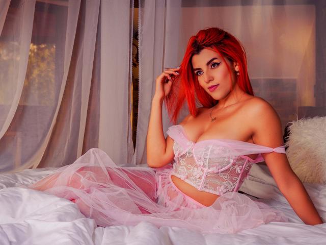 ZoeeMar with bright red hair, sitting on a bed in pink lace lingerie, showing her tits, with sheer fabric and a fluffy pillow. Her hand touches her hair.