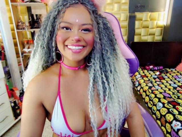 NashaJonny, met krullend, grijs tot wit haar, een roze choker en een witte bikini top die haar borsten laat zien, glimlacht naar de camera in een kleurrijke slaapkamer. Ze draagt roze oorbellen..