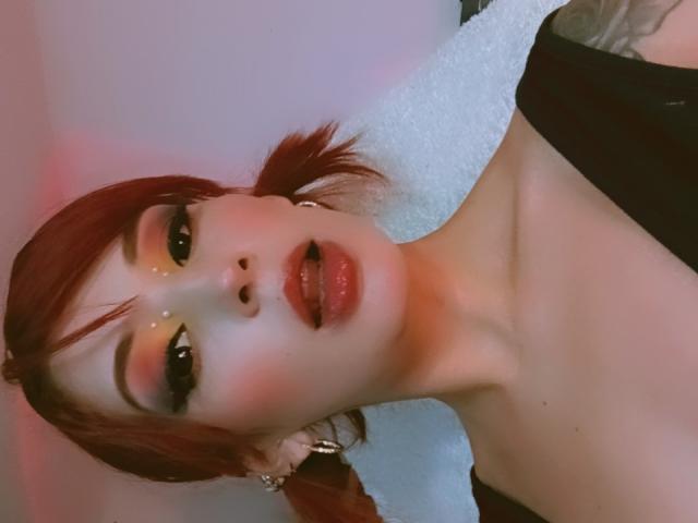 AriadnaFetish69 with red pigtails, glossy pink lips, and half-open mouth, wearing a black top, showing her bare shoulders and a white towel in the background.