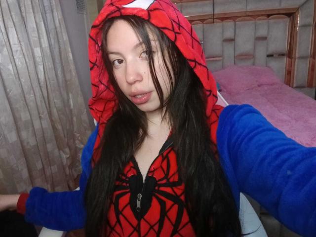 KattySnow, met lang zwart haar, draagt een rode en blauwe Spider-Man hoodie, heeft een lichte glimlach en haar armen zijn uitgestrekt. Op de achtergrond is een slaapkamer te zien, met een roze kussen..