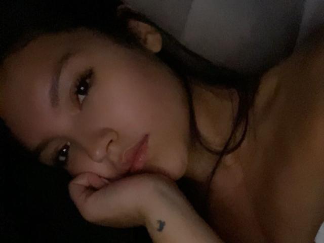 IssaPuasini lying down, head resting on hand, pouty lips, dark hair, brown eyes, slight smile, bare shoulders, small tattoo on hand, dim lighting, soft skin.
