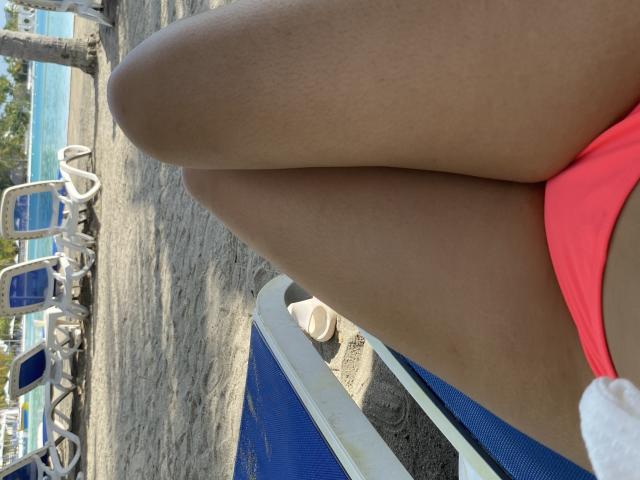 IssaPuasini's tan thighs with a bright pink bikini bottom, legs slightly parted, sitting on a blue chair by a pool, white sandals on the ground, sunny beach setting.