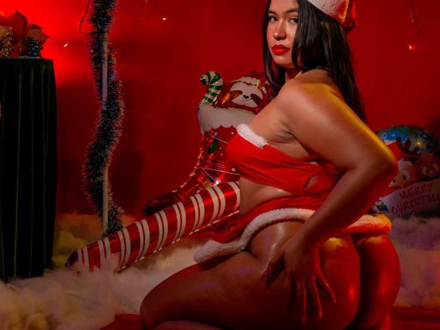 RossyGranger, curvy with red lipstick, wearing a Santa hat and red lingerie, has striped thigh-highs, exposing her big ass, and is holding a Santa face prop. Red lighting.