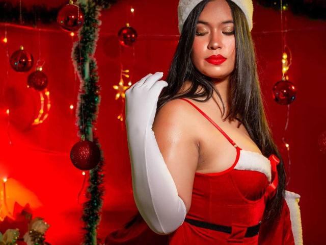 Rossy Granger mit langem, schwarzem Haar, rotem Lippenstift und weißen Handschuhen, trägt ein rotes Samt-Weihnachtskleid mit weißem Fellbesatz und einen weißen Weihnachtsmannhut. Sie hält ihre Schulter..