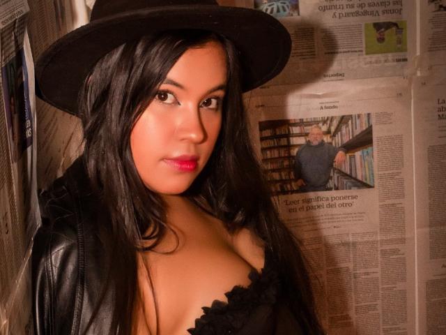 Rossy Granger, con i capelli neri, gli occhi marroni e le labbra rosa, indossa un cappello nero e una giacca di pelle, mostrando il décolleté con una maglia nera, e guarda di lato..