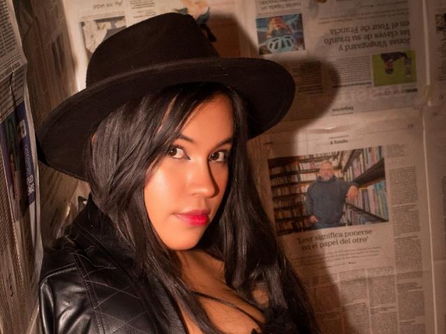 RossyGranger with black hat, long black hair, red lipstick, and black leather jacket, showing off her tits. Newspaper in background. Warm light. Sassy, sexy, confident.