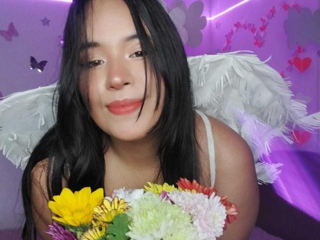 RossyGranger with black hair, angel wings, and red lipstick, holding colorful flowers, smiling slightly, in a purple-lit room. RossyGranger's medium tits visible through white top.