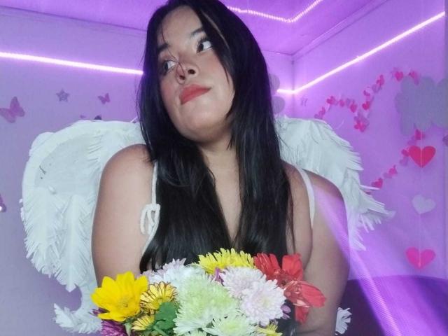 RossyGranger with black hair, red lipstick, white angel wings, holding colorful flower bouquet, purple LED strip lights, looking up with a slightly confused expression.