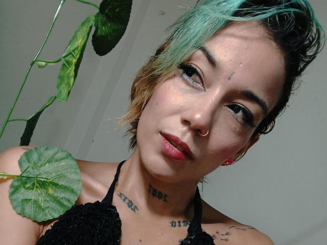 Veroo69 met turquoise en bruin haar, een neusring, tatoeages en roze lippenstift, draagt een zwarte kanten top en heeft een zachte glimlach. Op de achtergrond zijn groene plantenbladeren te zien..
