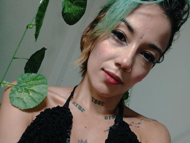 Veroo69 with short, green-blue, and brown hair, black lace bra, nose ring, tattoos on neck, and shoulder, smiling at camera, green plant leaves in background.