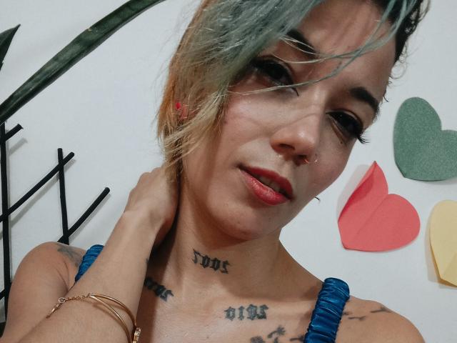 Veroo69 with blonde-green hair, nose ring, red lipstick, tattoos on neck and chest, blue straps, gold bracelet, hand on neck, smiling, heart decorations in background.