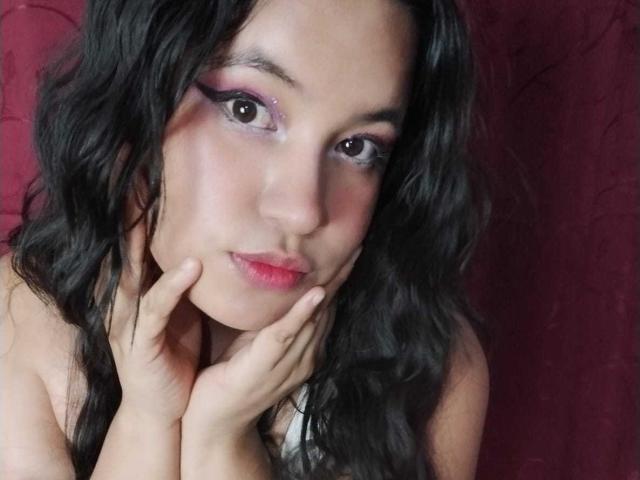 RossyGranger has wavy black hair, pink eyeshadow, and pink lipstick. She's touching her face with hands, showing off her shiny skin and pouty lips. Her ass is not visible.