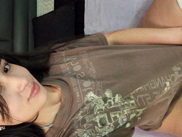 ValeriiNaughty with dark hair, light skin, and pink lips, wearing a brown graphic tee, lying on a bed, showing her bare thighs and a hint of her ass.