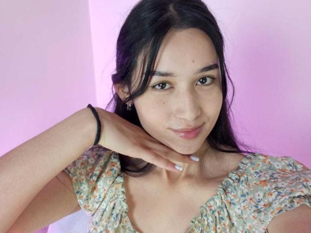 ValeriiNaughty has long black hair, brown eyes, light skin, small nose, subtle smile, wearing a floral dress, black wristband, and her hand rests on her neck.