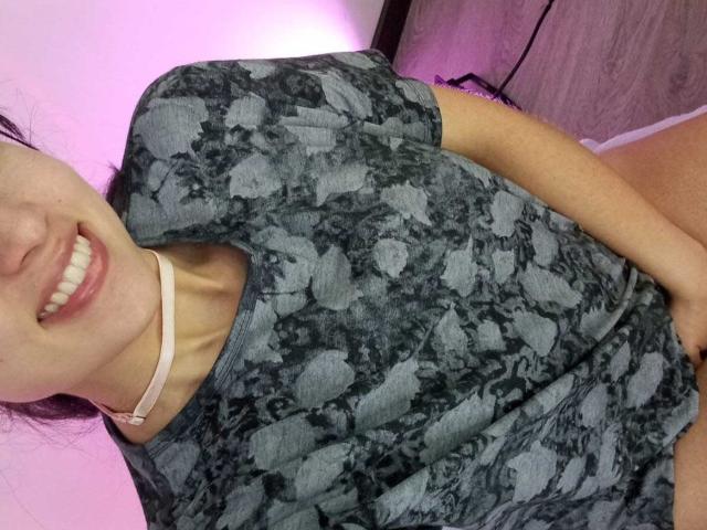ValeriiNaughty in a green and black camouflage t-shirt, smiling with white teeth, wearing a white choker, holding her crotch with her right hand, pink light in background.
