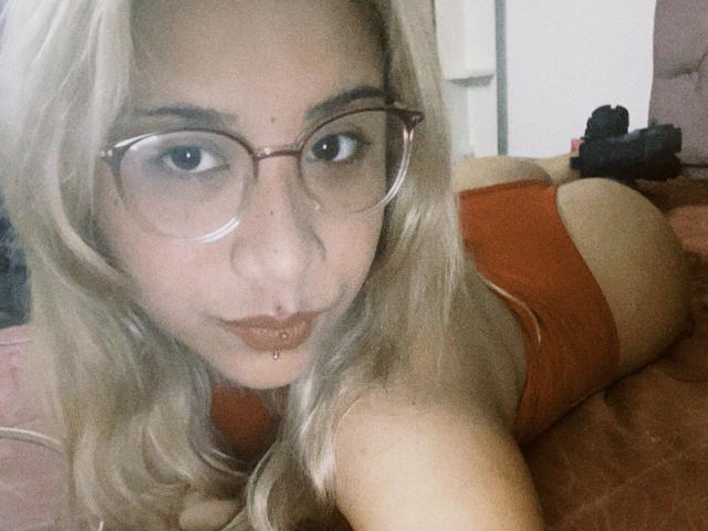 ExodiaKinky with blonde hair, glasses, and orange lingerie, lying on a bed, showing her ass, with a lip piercing and a slightly pouty expression.