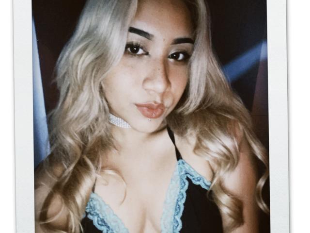 ExodiaKinky with blonde wavy hair, wearing a black and blue lace-trimmed top, choker, and lip piercing, looking at the camera with pouty lips.
