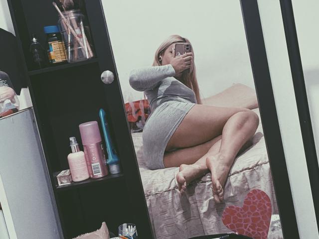ExodiaKinky in a gray long-sleeve dress, sitting on a bed, showing her ass and legs in a mirror selfie, with pink and blue sex toys on a shelf.