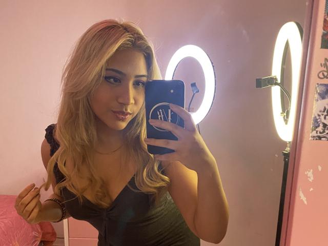 ExodiaKinky with blonde hair, wearing a black dress, taking a mirror selfie with a ring light in the background, holding a blue phone, showing her tits and a slight smile.
