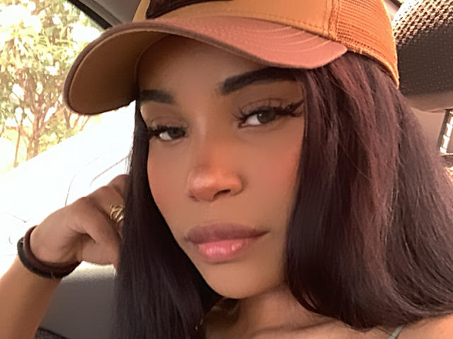 Clawdeenfoxy has dark brown hair, green eyes, and is wearing a brown cap, with a ring on her right hand and a black bracelet. She's looking at the camera.