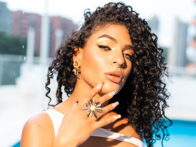 VioletHotX with curly black hair, golden eye makeup, shiny lips, white top, star earrings, silver ring, and hand touching her chin, standing by a blue pool.