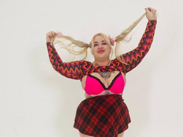 KateJen con due codini biondi, indossa un reggiseno rosa, maniche lunghe a zig-zag rosse e nere, e una gonna a quadri rossa, tiene i codini, mostrando i suoi seni e un tatuaggio sul petto..