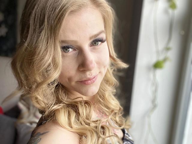 PixiFleur-hot has blonde curly hair, green eyes with heavy eyeliner, a slight smile, and a tattoo on her right shoulder, wearing a black top. She's near a window.