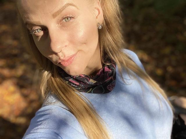 PixiFleur-hot with blonde hair, light skin, green eyes, subtle smile, small nose, wearing a blue sweater, black floral scarf, and diamond earrings, in sunlight.