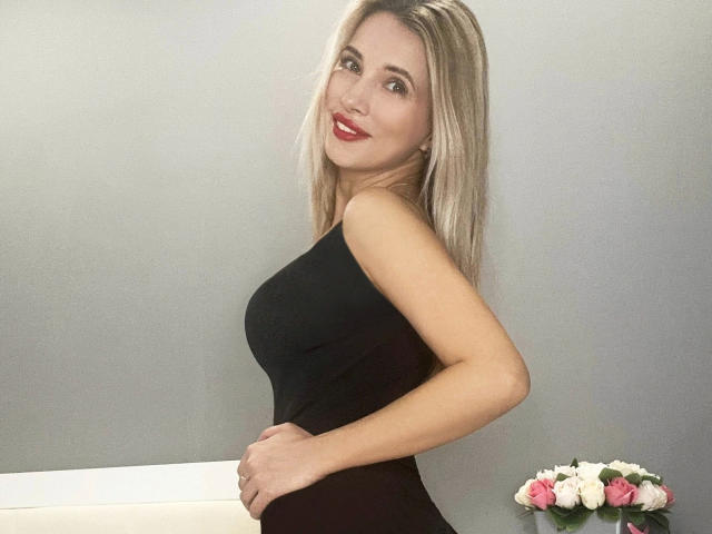 RutieGold-hot with blonde hair, red lipstick, black sleeveless dress, standing sideways, smiling, hand on hip, white couch, pink and white roses in the background.