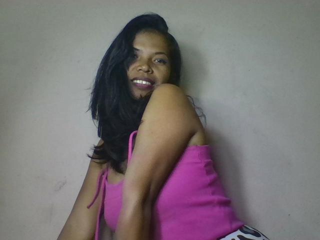 AlissaHott with dark skin, black wavy hair, pink tank top, smiling, looking over shoulder, showing her ass, against plain wall.