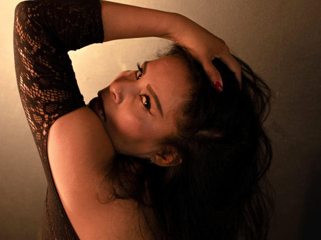 Anthoanela mit dunklem Haar, die ein schwarzes Spitzenoberteil trägt, hat ihre Hand in ihrem Haar, blickt mit verführerischen Augen in die Kamera, hat rote Nägel und einen sexy, selbstbewussten Ausdruck..