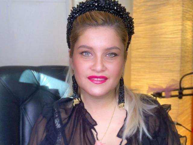 KillerSmilexxx-hot has pink lipstick, black sheer top, black bead headpiece, and black bead earrings. She's sitting in a black leather chair, looking at the camera.