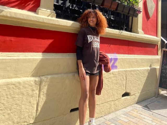 Cinthia Walker, avec ses cheveux roux bouclés, portant une chemise marron foncé ample, un short noir et des chaussettes blanches, appuyée contre un mur jaune et rouge, tenant une veste marron..