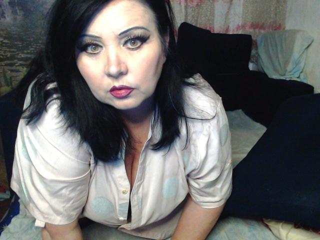KamilaDream with dark hair, heavy eyeliner, and red lipstick, looking up with pouty lips, wearing a white button-up shirt with blue circles, showing her big tits.