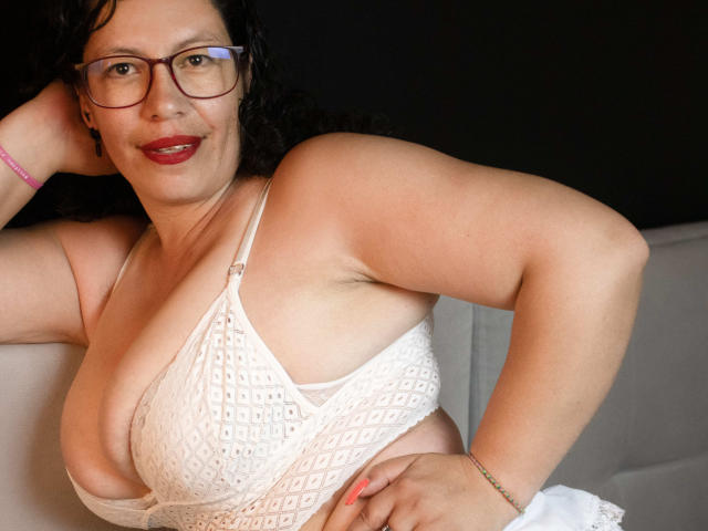 ZalyMature with glasses, red lipstick, and curly black hair, wearing a white patterned bra that reveals her large, soft tits. She's smiling, resting her arm on a surface.
