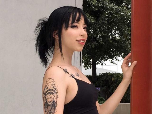Arieyaara with black hair in a ponytail, wearing a black tank top, showing a tattoo on her right arm, smiling, standing next to a red pillar, outdoors.