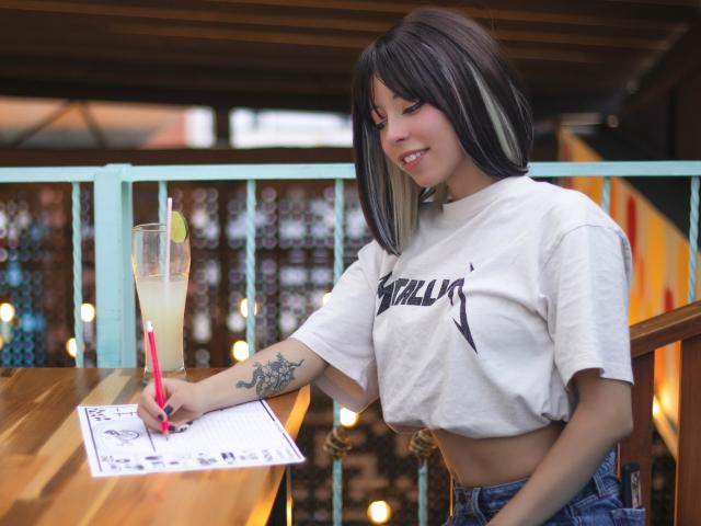 Arieyaara with dark brown and white hair, wearing a white crop top and blue jeans, sits at a wooden table, drawing with a red pen, next to a tall drink.