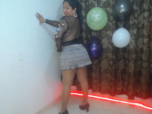 CatalinaJH with black sheer top, silver sequins, grey plaid skirt, black high heels, standing against white wall, back to camera, red LED strip, balloons, brown curtains.