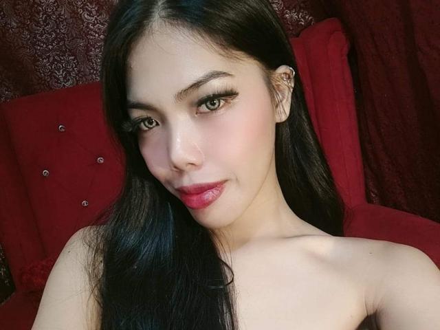 HaileeHarper with long black hair, pink lipstick, and subtle makeup, showing her bare shoulders and a small earring, smiling softly at the camera.