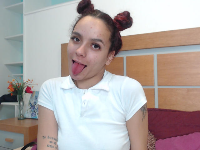 MaiaPark with red double buns, white polo, tongue out, tattoos on right arm, sitting on bed with wooden headboard, pink and maroon pillows, light skin, brown eyes.