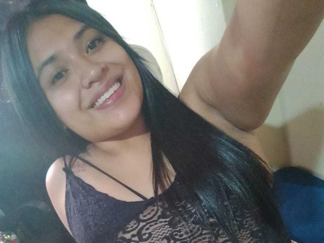 AmyWhin, smiling with black lace top, showing tits, long straight black hair, brown eyes, tan skin, white teeth, raised arm, in a selfie.
