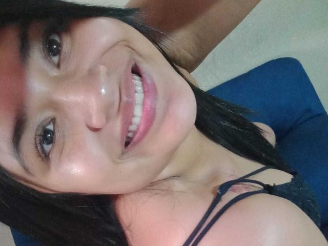 AmyWhin with dark straight hair, smiling with white teeth, wearing black lace top, showing small tits, tan skin, and brown eyes, taking a selfie.
