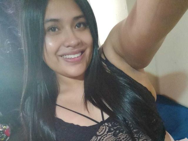 AmyWhin with long black hair, smiling, wearing a black lace top, showing her tits, holding up a selfie with one arm. Her skin is tan, and she's looking at the camera.