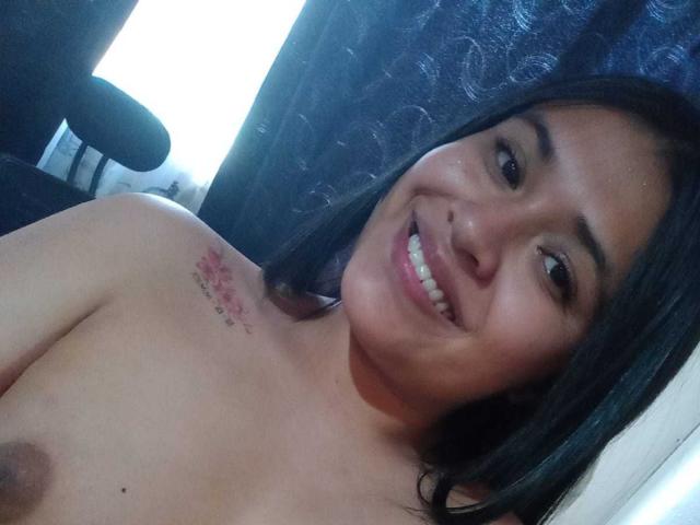AmyWhin has medium brown skin, black hair, smiling with teeth showing, topless with small tits and a pink tattoo on her right shoulder. Her nipples are visible.