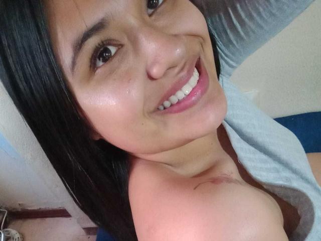 AmyWhin, smiling with dark eyes, bare shoulders, and black hair, looking at camera, showing small tits, gray shirt, and pink lips, head tilted, arm raised.