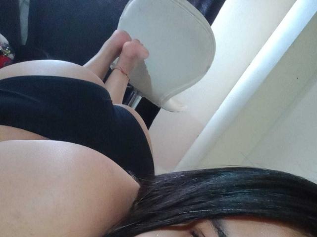 AmyWhin lying on her stomach, black panties, bare butt up, feet on chair, long dark hair, visible eye, soft skin, red ankle bracelet, white chair.