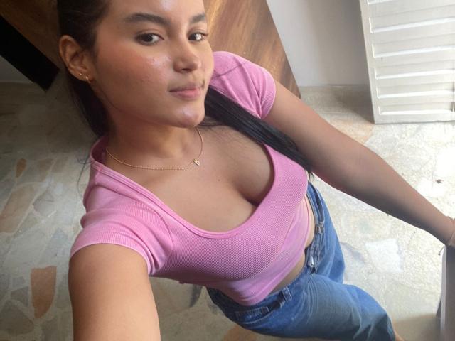 MiiaPalmer with tan skin, black hair in a ponytail, wearing a pink tight shirt showing her big boobs and blue jeans, standing in a room with a stone floor.