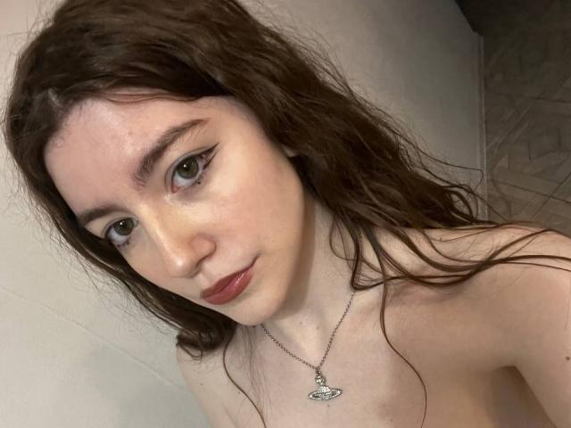 VinnaSkyHott has pale skin, green eyes with winged eyeliner, red lips, long brown wavy hair, and wears a necklace with a small pendant, topless, showing her bare shoulders.