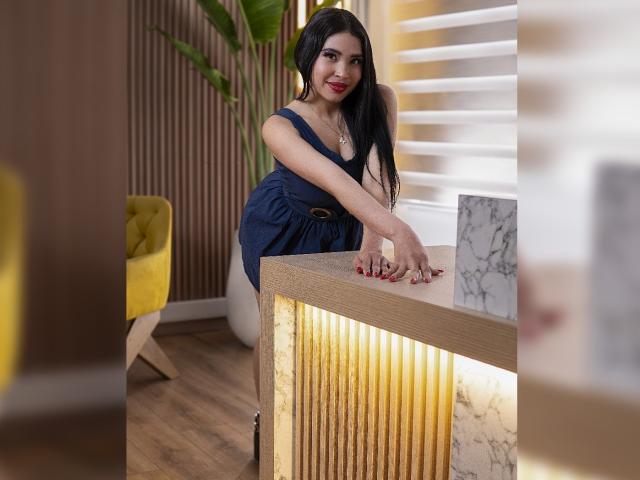 OliviGrace with long black hair, red lipstick, and a navy dress leaning on a wooden counter with marble accents, smiling, hands on the counter, wooden floor, yellow chair.
