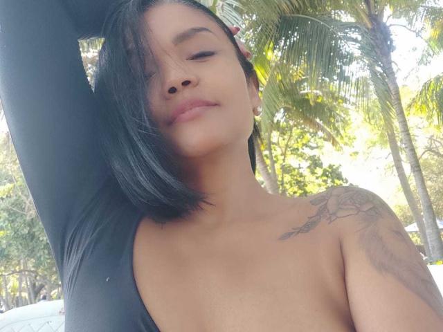 ExoticLyla met zwart haar, ogen gesloten, lachend, gekleed in een zwarte top, waarbij haar kleine borsten en een tatoeage op haar rechter schouder zichtbaar zijn, arm omhoog, staand buiten, in het zonlicht..